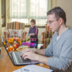 Father working hard in home office with notebook and his boring son playing on the background